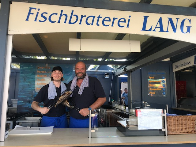 Zwei Mitarbeiter der Fischbraterei Lang präsentieren frisch gegrillten Fisch
